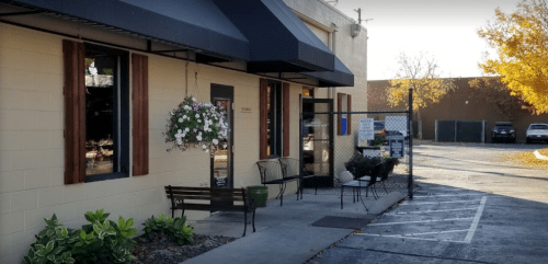 A small building with a black awning, hanging flower baskets, and a fenced area in a parking lot with autumn trees.