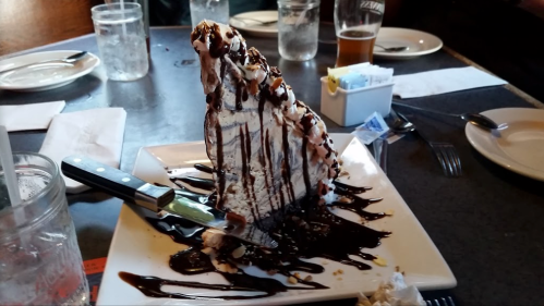 A large slice of chocolate cake drizzled with chocolate sauce on a white plate, with a knife beside it.