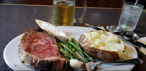 A plate with prime rib, asparagus, a baked potato, and a slice of bread, accompanied by a beer and water.