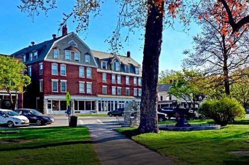 Historic brick hotel with large windows, surrounded by trees and parked cars on a sunny day.