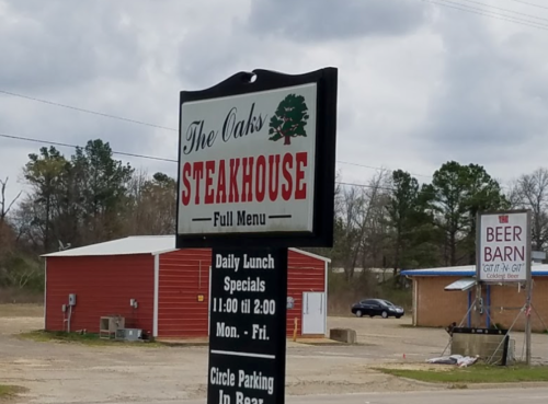 Sign for The Oaks Steakhouse with "Full Menu" and daily lunch specials, alongside a Beer Barn sign in the background.