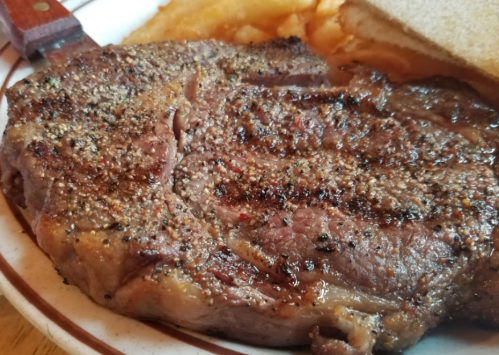 A close-up of a seasoned, grilled steak on a plate with fries and a slice of bread.