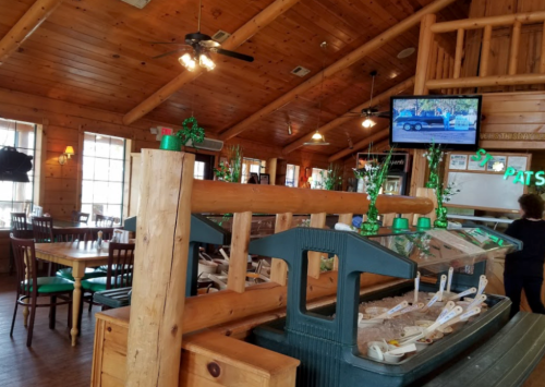 Interior of a rustic restaurant with wooden decor, tables, and a TV displaying a vehicle. St. Patrick's Day decorations visible.