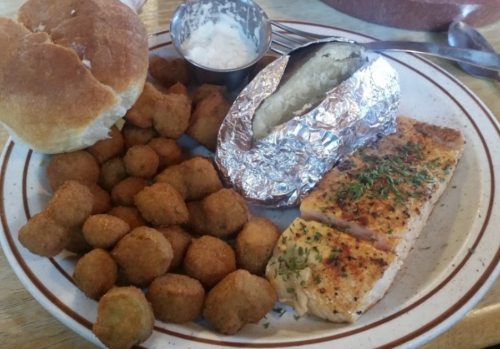 A plate featuring baked fish, a baked potato in foil, fried okra, and a roll with a side of dipping sauce.