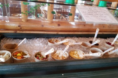 A display of various toppings in cups on ice, with wooden spoons, set in a rustic restaurant environment.