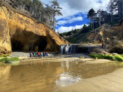 A scenic beach with a waterfall, rocky cliffs, and a group of people exploring the area under a partly cloudy sky.