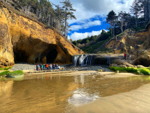 A scenic beach with a waterfall, rocky cliffs, and a group of people exploring the area under a blue sky.