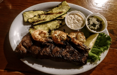 A plate featuring grilled steak, shrimp, zucchini, and dipping sauces, garnished with lettuce and a lemon wedge.