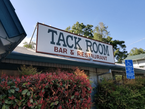 Sign for "Tack Room Bar & Restaurant" with greenery and trees in the background.
