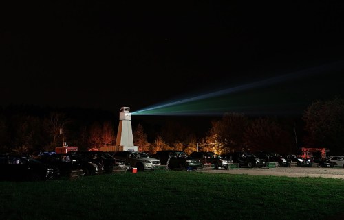 A lighthouse beam illuminates a dark parking lot filled with cars at night, surrounded by trees.