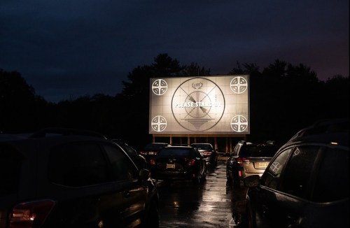 A drive-in movie screen displays a "Please Stand By" message at night, surrounded by parked cars.