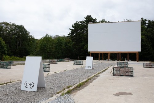 An empty outdoor movie screen with gravel ground and signage, surrounded by trees under a cloudy sky.