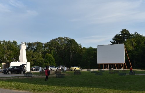 A drive-in movie setup with a large blank screen, cars parked, and people near a concession area in a green field.