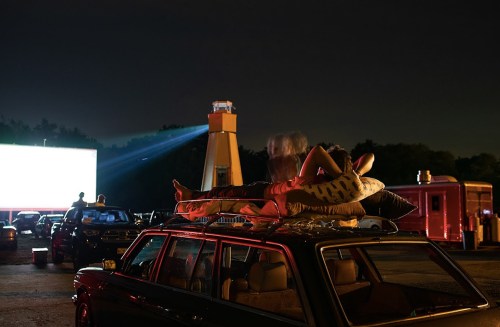 A person relaxes on pillows atop a car at a drive-in movie, with a large screen and a lighthouse in the background.