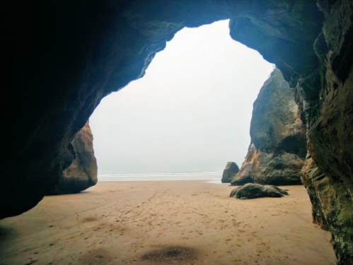 View from inside a rocky cave, looking out at a sandy beach and ocean under a cloudy sky.