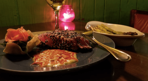 A plate of sesame-crusted tuna with sauce, garnished with vegetables, alongside asparagus and a candlelit ambiance.
