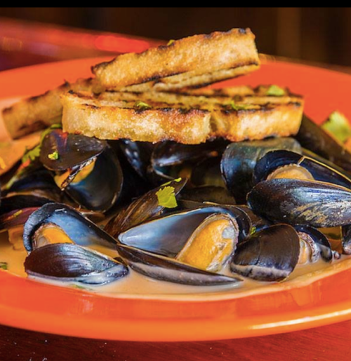 A vibrant orange plate with mussels in a creamy sauce, topped with grilled bread and garnished with herbs.