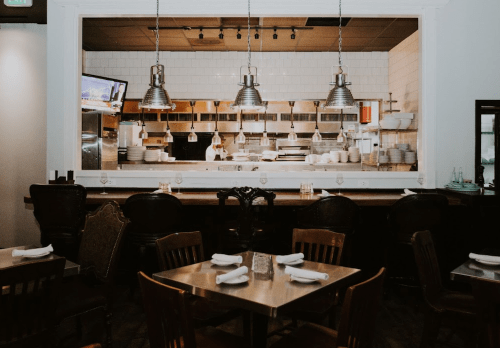A cozy restaurant interior with wooden tables, chairs, and an open kitchen visible in the background.