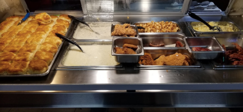 A buffet spread featuring biscuits, gravy, fried chicken, potatoes, and various meats in metal trays.