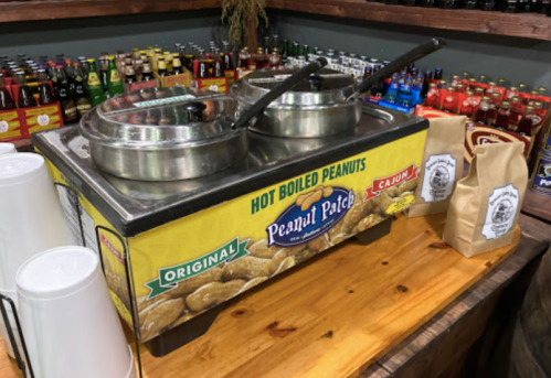 A hot boiled peanuts stand with two pots, cups, and bags of peanuts on a wooden counter, surrounded by drinks.