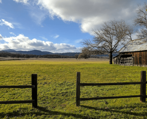 A scenic landscape featuring a grassy field, a bare tree, a rustic barn, and distant mountains under a partly cloudy sky.