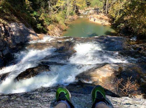 A view from above a waterfall, with feet in green shoes at the edge and a serene river below surrounded by trees.