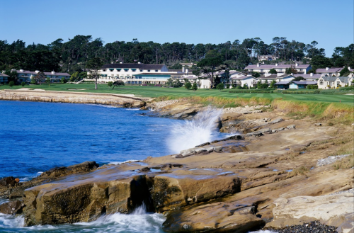 A scenic view of a coastline with waves crashing against rocks, overlooking a golf course and lush greenery in the background.