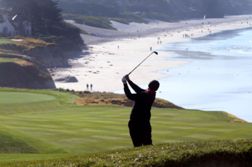 A golfer swings on a lush green course overlooking a sandy beach and ocean, with people walking along the shore.