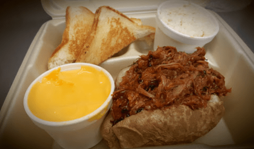 A takeout container with pulled pork on a bun, cheese sauce, coleslaw, and toasted bread slices.