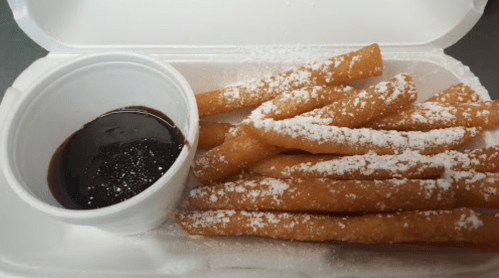 A takeout container with fried dough sticks dusted with powdered sugar and a small cup of chocolate sauce.