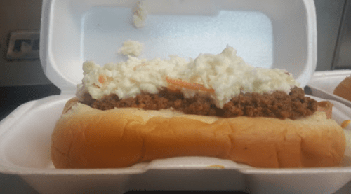 A hot dog in a foam container topped with seasoned ground meat and a creamy coleslaw mixture.