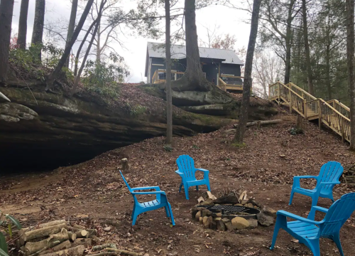 A cozy outdoor scene with blue chairs around a fire pit, a wooden cabin on a hill, and trees in a wooded area.