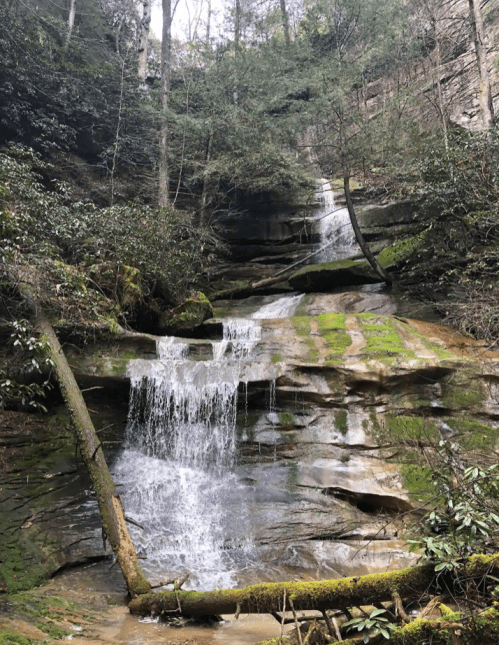 A serene waterfall cascades over moss-covered rocks, surrounded by lush greenery and steep cliffs.