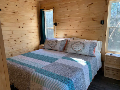Cozy bedroom with a wooden interior, featuring a double bed with a patterned quilt and large windows for natural light.