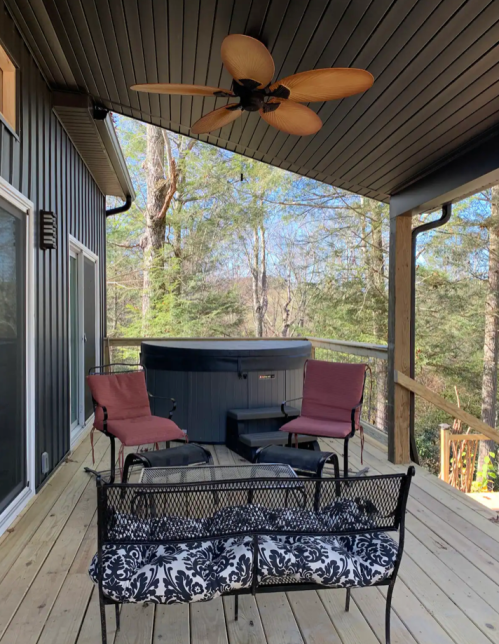 A wooden deck with a hot tub, two chairs, and a bench, surrounded by trees and a clear blue sky.
