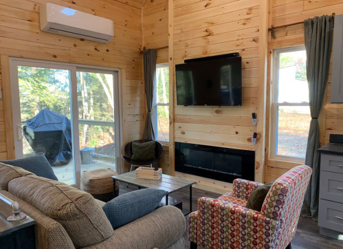 Cozy living room with wooden walls, a TV, fireplace, and large windows overlooking a wooded area.