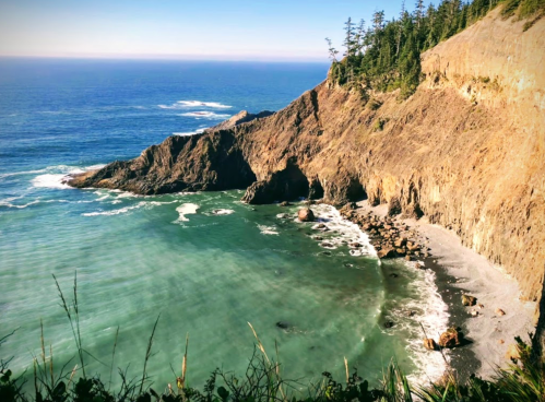 A scenic coastal view featuring rocky cliffs, a sandy beach, and calm blue waters under a clear sky.
