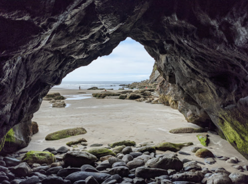 View from inside a rocky cave, looking out at a sandy beach and ocean under a cloudy sky.