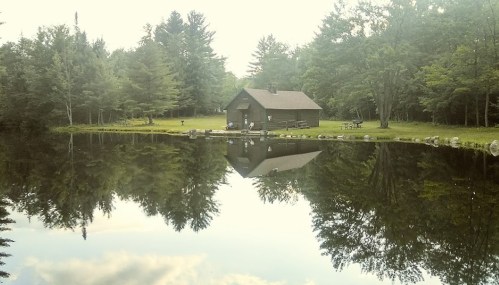 A serene cabin by a calm pond, surrounded by trees, reflecting the peaceful landscape.