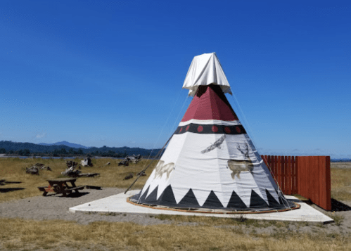 A colorful teepee stands on a grassy area with mountains in the background under a clear blue sky.