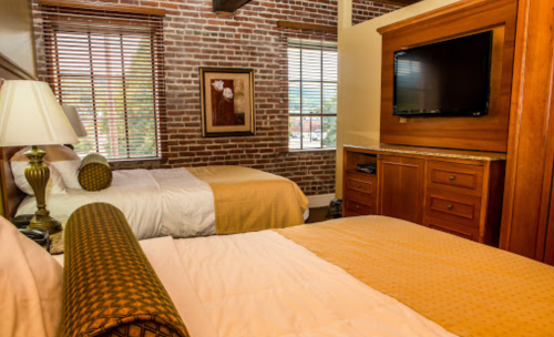 Cozy hotel room with two beds, brick walls, wooden furniture, and a flat-screen TV. Natural light from window.