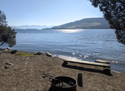 A serene lakeside view with mountains in the background, featuring a fire pit and a wooden bench on the shore.