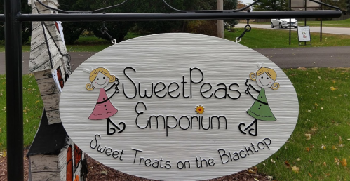 Sign for "Sweet Peas Emporium" featuring playful illustrations of girls, advertising sweet treats on the blacktop.