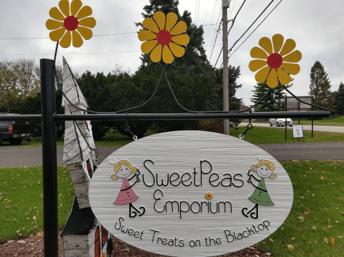 Sign for "SweetPeas Emporium" featuring flowers and playful illustrations, advertising sweet treats on the blacktop.
