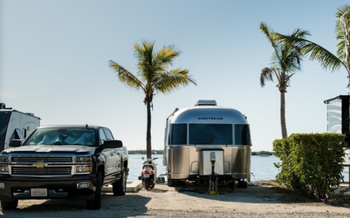 A silver Airstream trailer parked by the water, with a black truck and palm trees in the background.
