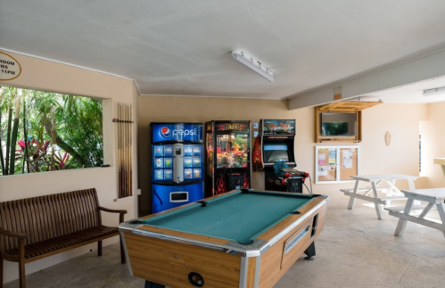 A recreational room featuring a pool table, vending machines, and seating in a bright, tropical setting.