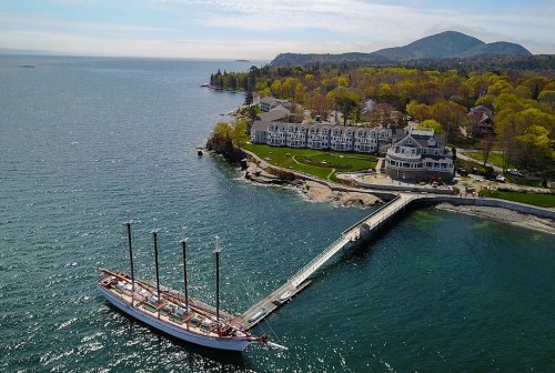 Aerial view of a docked tall ship near a waterfront hotel and lush green hills under a clear sky.