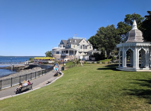 A scenic waterfront view featuring a historic house, gazebo, and people enjoying a sunny day by the water.
