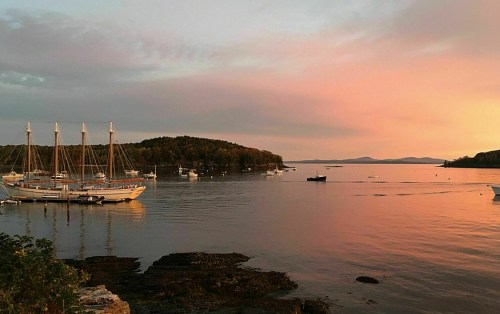 A serene sunset over a calm bay with sailboats and an island in the distance, reflecting warm colors on the water.