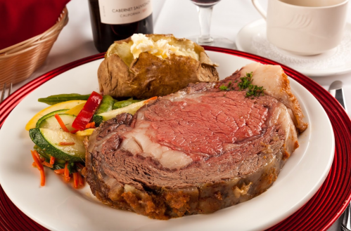 A plate featuring a juicy prime rib, baked potato, and mixed vegetables, with a glass of red wine in the background.